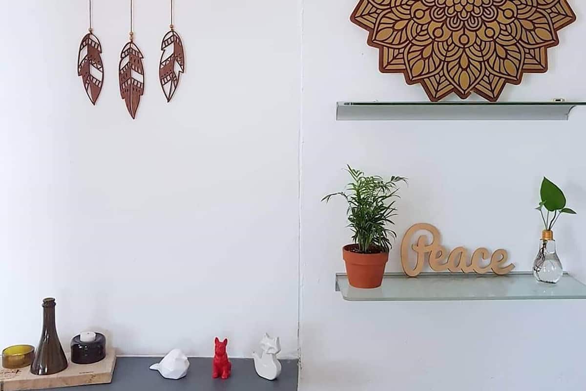 Una pared con un atrapasueños y una decoración de mandala. Abajo, hay estantes con una planta en maceta, un cartel con la palabra 'peace' y varios objetos decorativos pequeños.