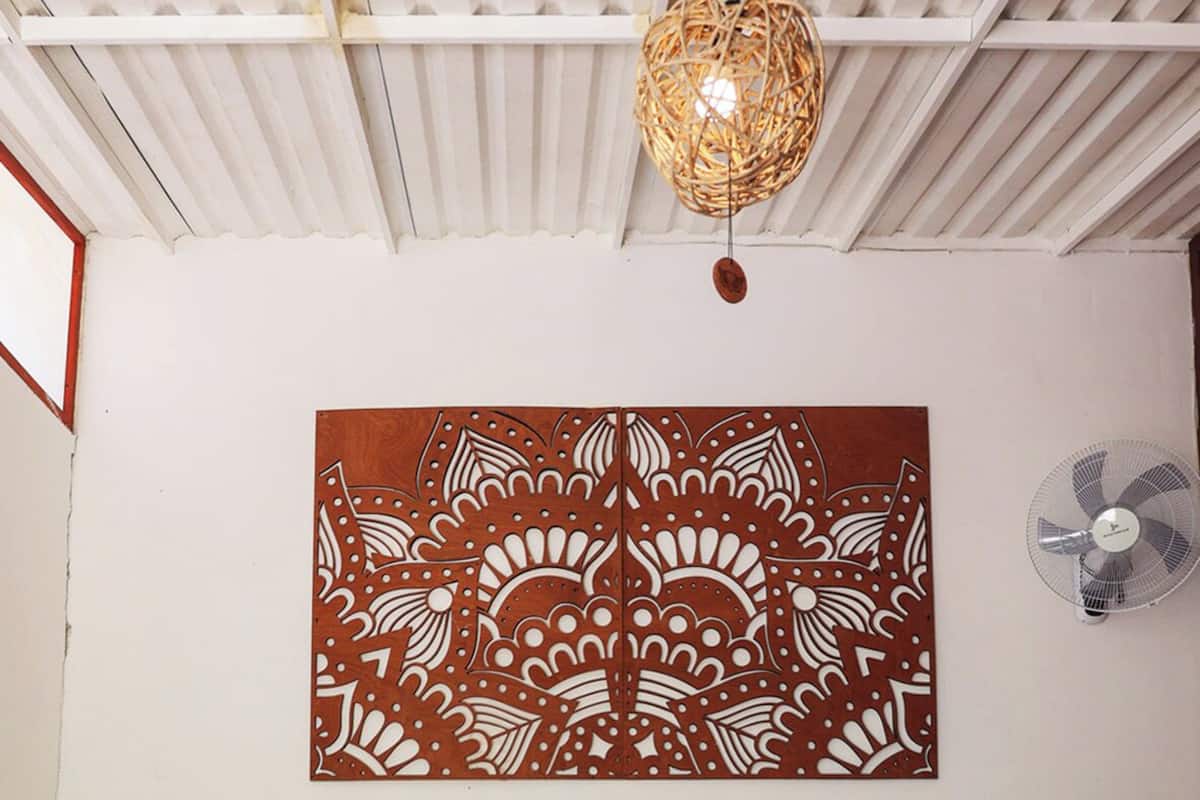 Una habitación con dos camas, un tapiz tejido, un adorno en la pared, un ventilador y un estante pequeño con una planta.