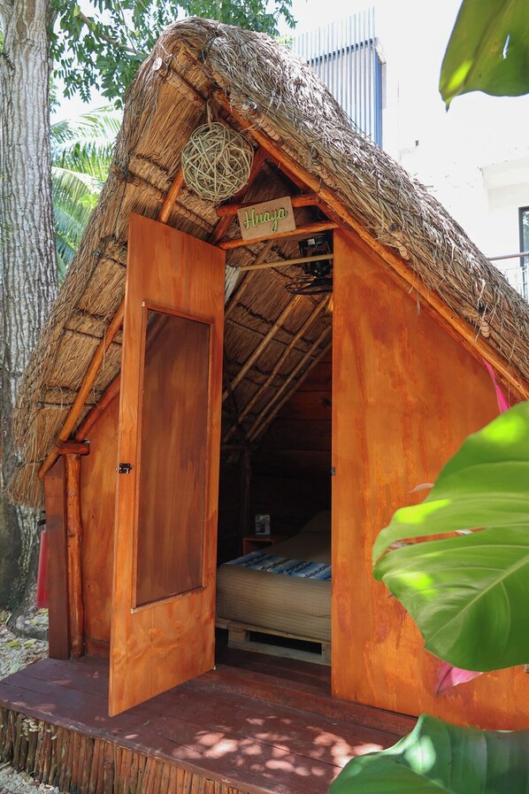 A thatched-roof hut with a wooden door labeled "Hriaya," a bed inside, and a small table. - El Búho Eco-Cabañas Hostal. (Bacalar)