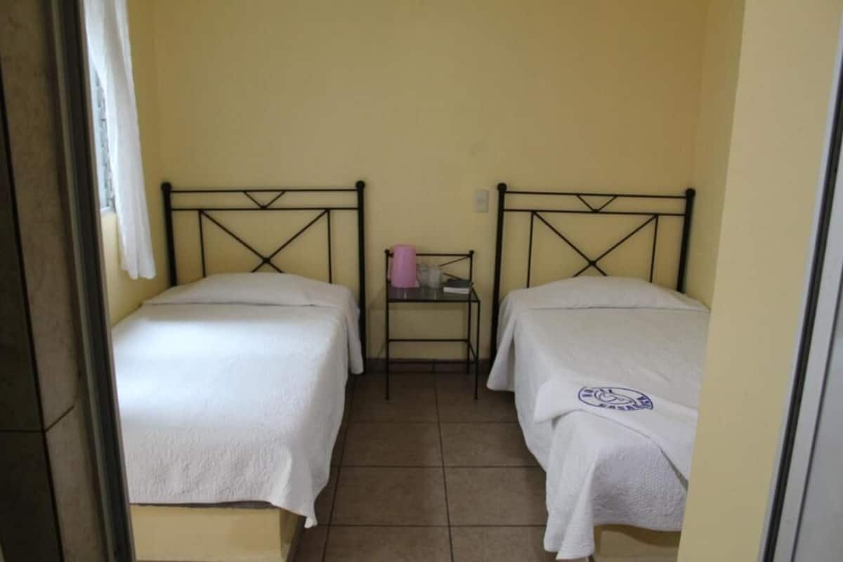 A narrow room with two single beds, each with a white bedspread and a metal frame. There is a small table with a pink object on it, and the walls are painted a light yellow.