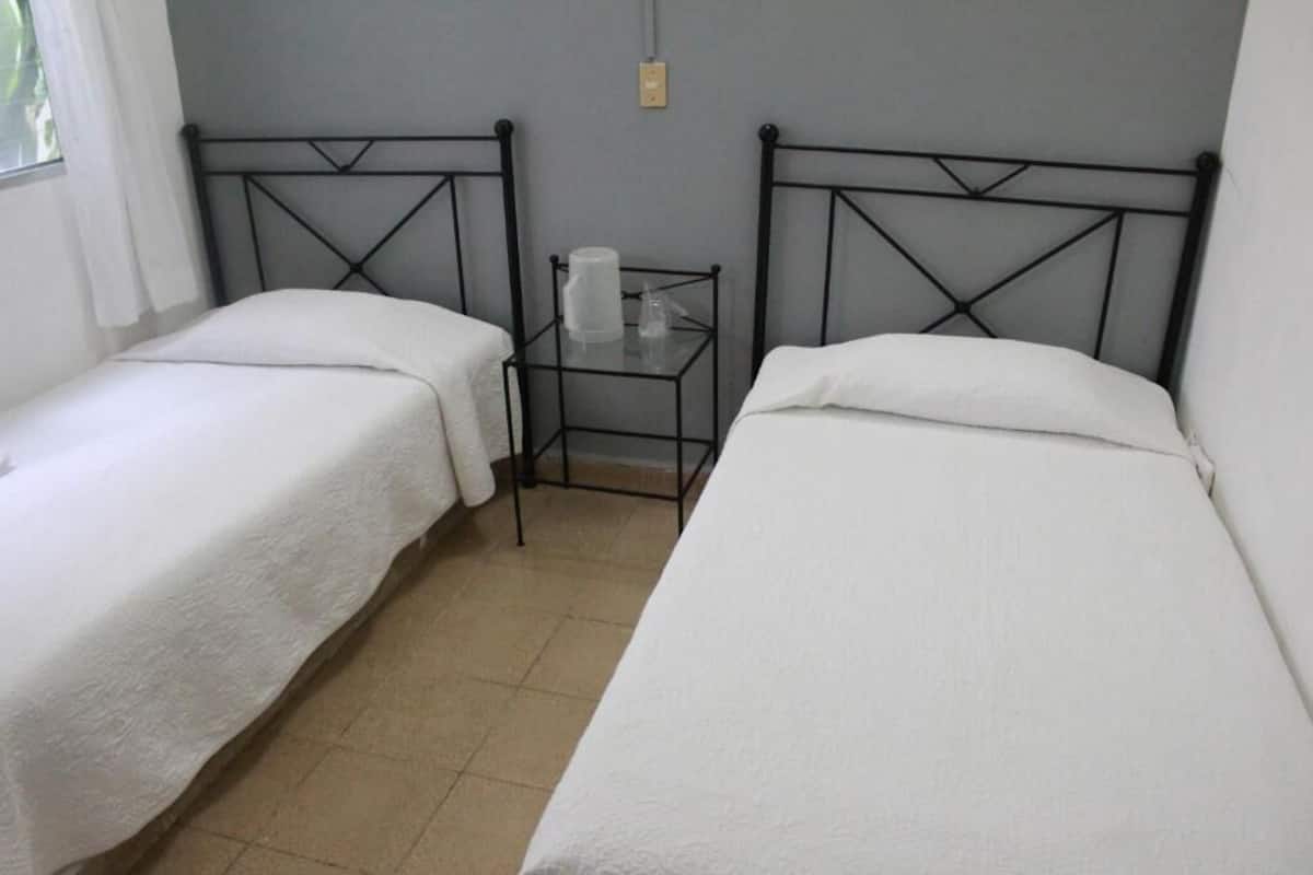 A room with two single beds, a small table with a cup and a tissue, and a plain wall with a light switch.