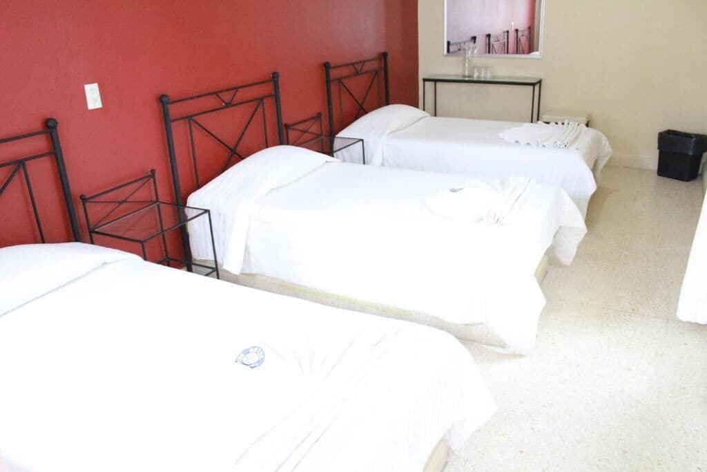 Habitación con tres camas individuales, cada una con estructura metálica y ropa blanca. Hay una mesita pequeña con un espejo y algunos objetos encima. Las paredes son rojas y el suelo es de color claro.