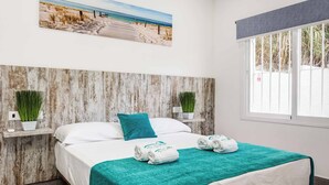 A bedroom with a wooden headboard, a bed with white linens, teal throw pillows, and towels. There is a painting of a beach scene on the wall, and a window with a view of greenery. - Villa Conchita Torremolinos (Torremolinos)