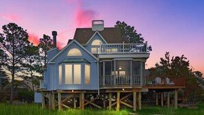 A two-story house with a balcony and a covered porch, elevated on stilts. - Blue Pointe Bayside | Cul-de-Sac Waterfront w/ 4 Decks, Screened Porch, Sunsets & Bay Views (Chincoteague Island)
