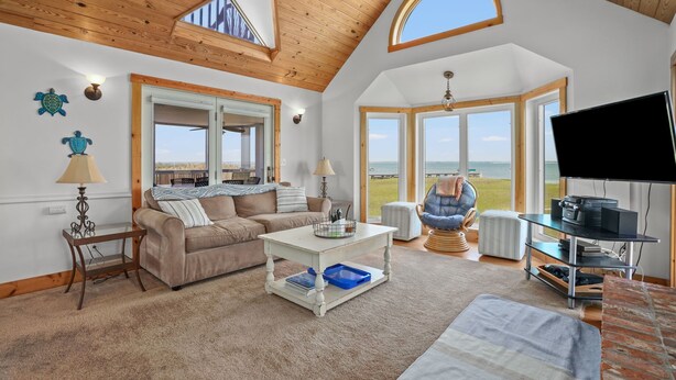 A spacious living room with a high ceiling, wooden beams, a large window overlooking a scenic view, a comfortable sofa, a coffee table, and a television. - Blue Pointe Bayside | Cul-de-Sac Waterfront w/ 4 Decks, Screened Porch, Sunsets & Bay Views (Chincoteague Island)
