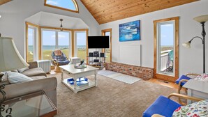 A spacious living room with a large window offering a view of the beach, a flat-screen TV mounted on the wall, and a cozy seating area with a fireplace. - Blue Pointe Bayside | Cul-de-Sac Waterfront w/ 4 Decks, Screened Porch, Sunsets & Bay Views (Chincoteague Island)