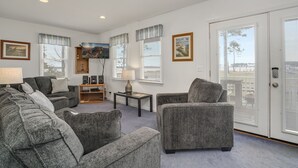 A living room with grey sofas, a coffee table, and a TV. There are framed pictures on the wall and a door with a view. - Channel Lookout | Memorial Park Water Views w/ Private Deck, Assateague Channel Scenery & Fishing (Chincoteague Island)