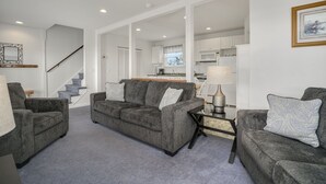 A living room with two grey sofas, a coffee table, and a lamp. A staircase leads to a kitchen area. - Channel Lookout | Memorial Park Water Views w/ Private Deck, Assateague Channel Scenery & Fishing (Chincoteague Island)