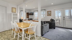A dining area with a wooden table and white chairs, a grey sofa, and a kitchen in the background. - Channel Lookout | Memorial Park Water Views w/ Private Deck, Assateague Channel Scenery & Fishing (Chincoteague Island)