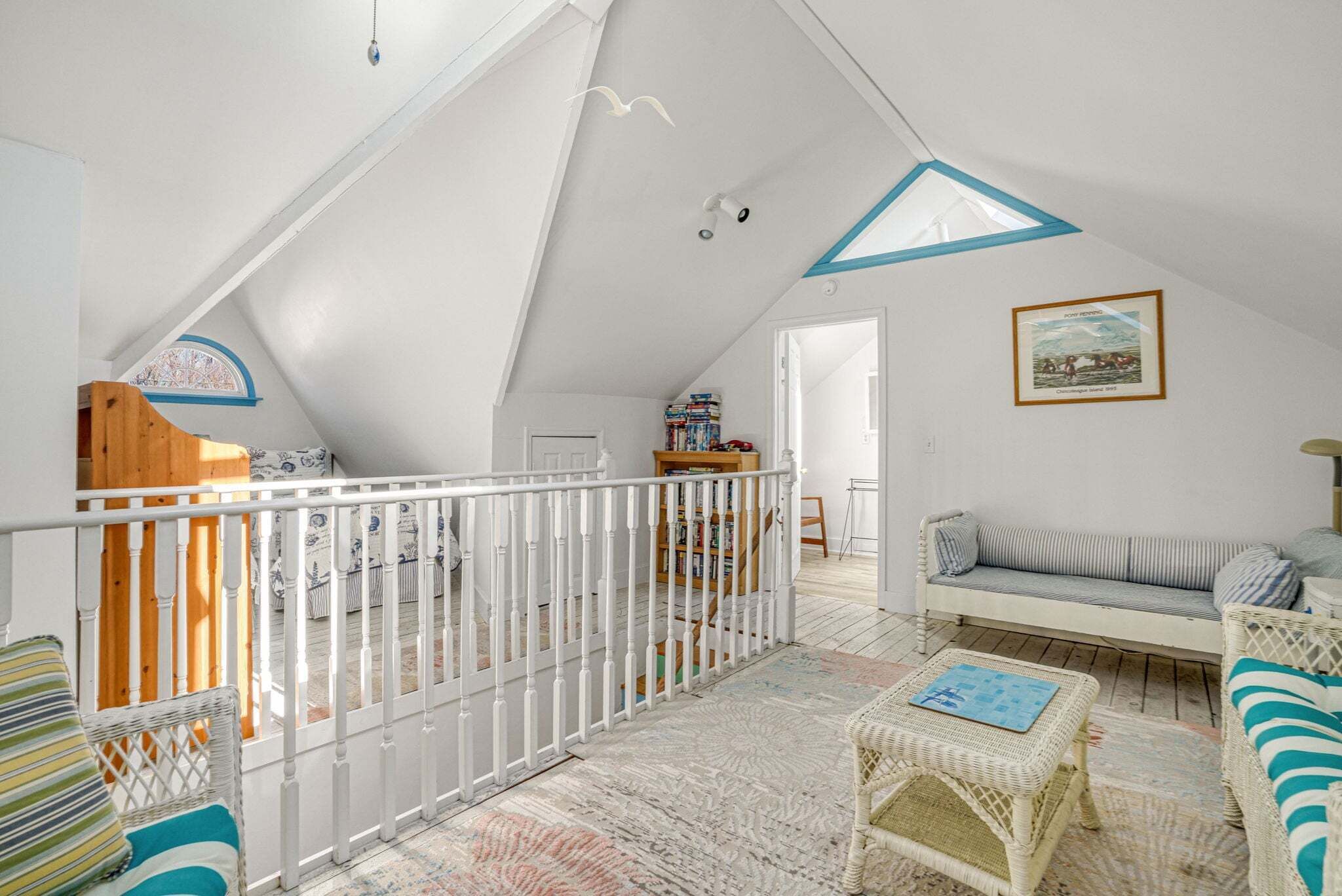 Una camera da letto mansardata con soffitto spiovente, ringhiera bianca, scala in legno, un divano, un tavolino e una libreria.