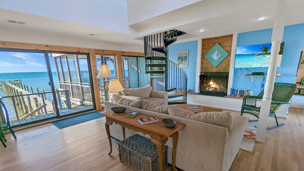 A spacious living area with a large window overlooking a wooden deck and the sea. A fireplace and bookshelves are visible in the background. - Grey Heron | Waterfront Home w/ Dock, Kayak Launch, Screened Porch, Epic Sunsets & Pet Friendly (Chincoteague Island)