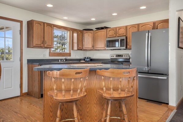 A kitchen with wooden cabinets, a stainless steel refrigerator, a microwave, and two wooden bar stools.