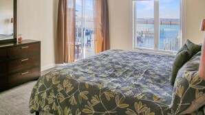 A bedroom with a large bed, a dresser, and a window with a view of the water. - Sunset Bay Villa 212 | Waterfront Balcony + Pool + Walk to Downtown + Pet Friendly (Chincoteague Island)