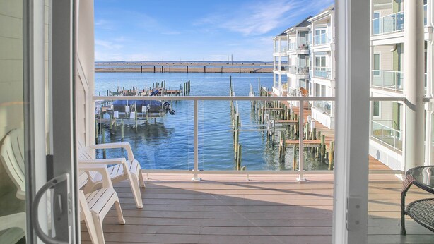 A balcony with a view of a dock and water, featuring white chairs and a glass railing. - Sunset Bay Villa 212 | Waterfront Balcony + Pool + Walk to Downtown + Pet Friendly (Chincoteague Island)