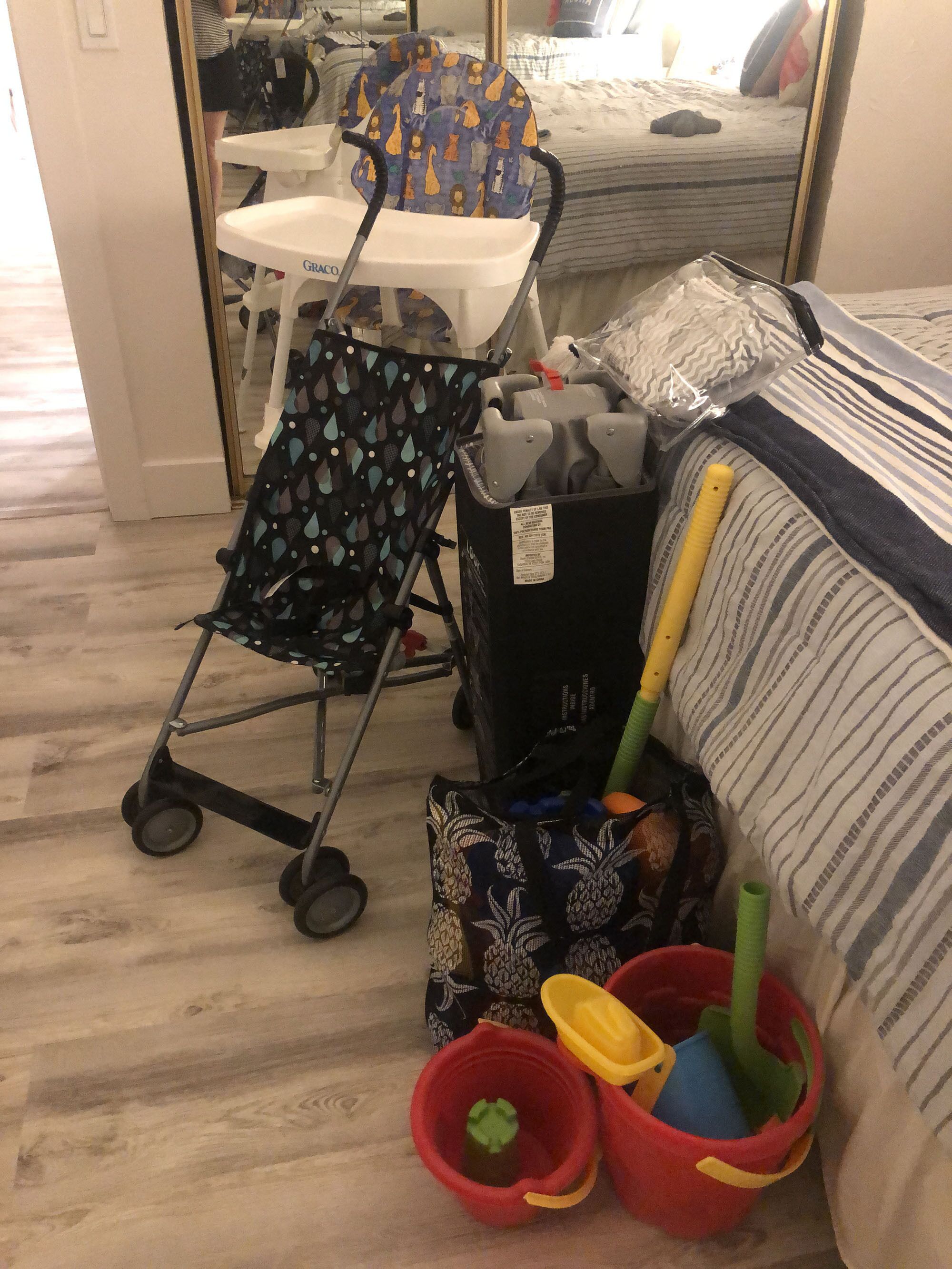 Una habitación con una cama, un cochecito, un cubo de basura y un cubo rojo con juguetes.