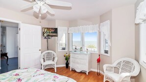 Un dormitorio con cama, dos sillas blancas de mimbre, cómoda, televisor y ventana con vistas.