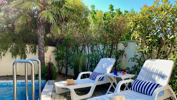 Un área junto a la piscina con dos sillones blancos, una escalera de metal y suelo de baldosas.