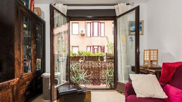 Un salon chaleureux avec un canapé rouge, un meuble en bois et une porte vitrée donnant sur un balcon agrémenté de plantes et offrant une vue sur un immeuble.