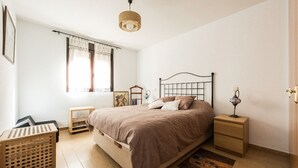Une chambre à coucher avec un lit, des tables de chevet, une fenêtre avec des rideaux, un luminaire suspendu et un plancher en bois.