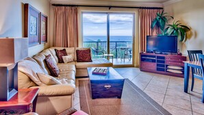 A living room with a sofa, coffee table, TV, and a balcony with a view. - 5th floor, Gulf views, 2 Pools, Free Beach Service, Garage Parking (Miramar Beach)