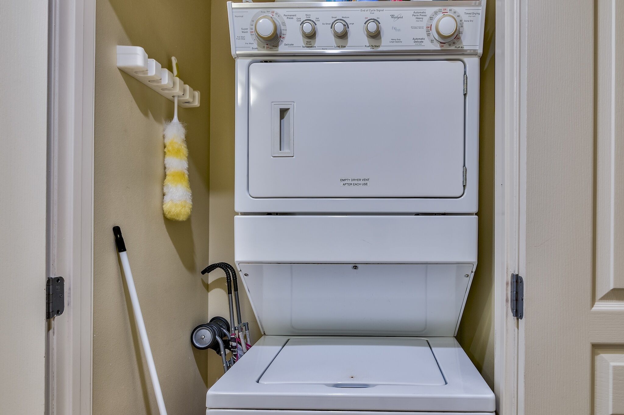 Una lavadora y secadora apiladas de color blanco, con una rejilla de ventilación en la parte superior, un trapeador y una escoba apoyados en la pared.