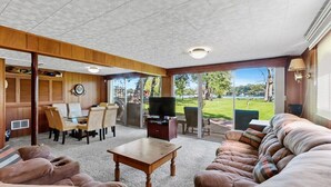 A spacious living room with a large couch, a coffee table, and a TV. There is a dining area with chairs and a table, and a view of a grassy area outside. - Lake Benedict Lakefront Home with Fire Pit Large Yard Kayak Friendly (Genoa City)