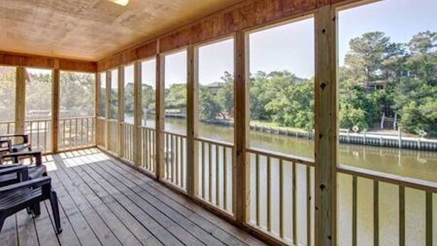 Un espace extérieur abrité, doté d’une terrasse en bois, de grandes fenêtres et offrant une vue sur une rivière et des arbres.