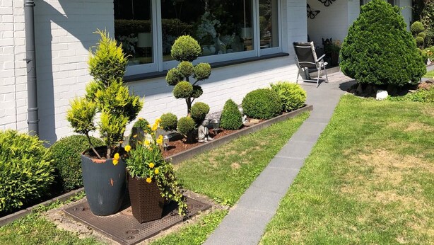 Une maison à deux étages, avec un toit en tuiles, des murs extérieurs blancs et un porche à l’avant. On y trouve des plantes en pot et un petit jardin agrémenté de divers arbustes et d’un arbre.