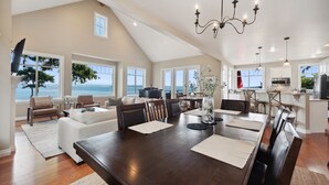 A spacious living room with a large dining table, comfortable seating, and a view of the ocean.