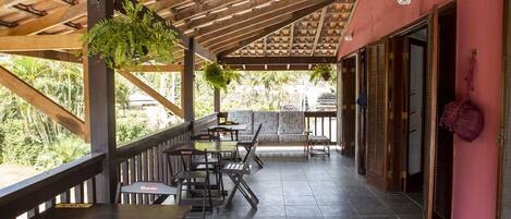 Un espace extérieur aménagé en terrasse, avec des tables et des chaises en bois, une fougère suspendue et une vue sur la verdure environnante.