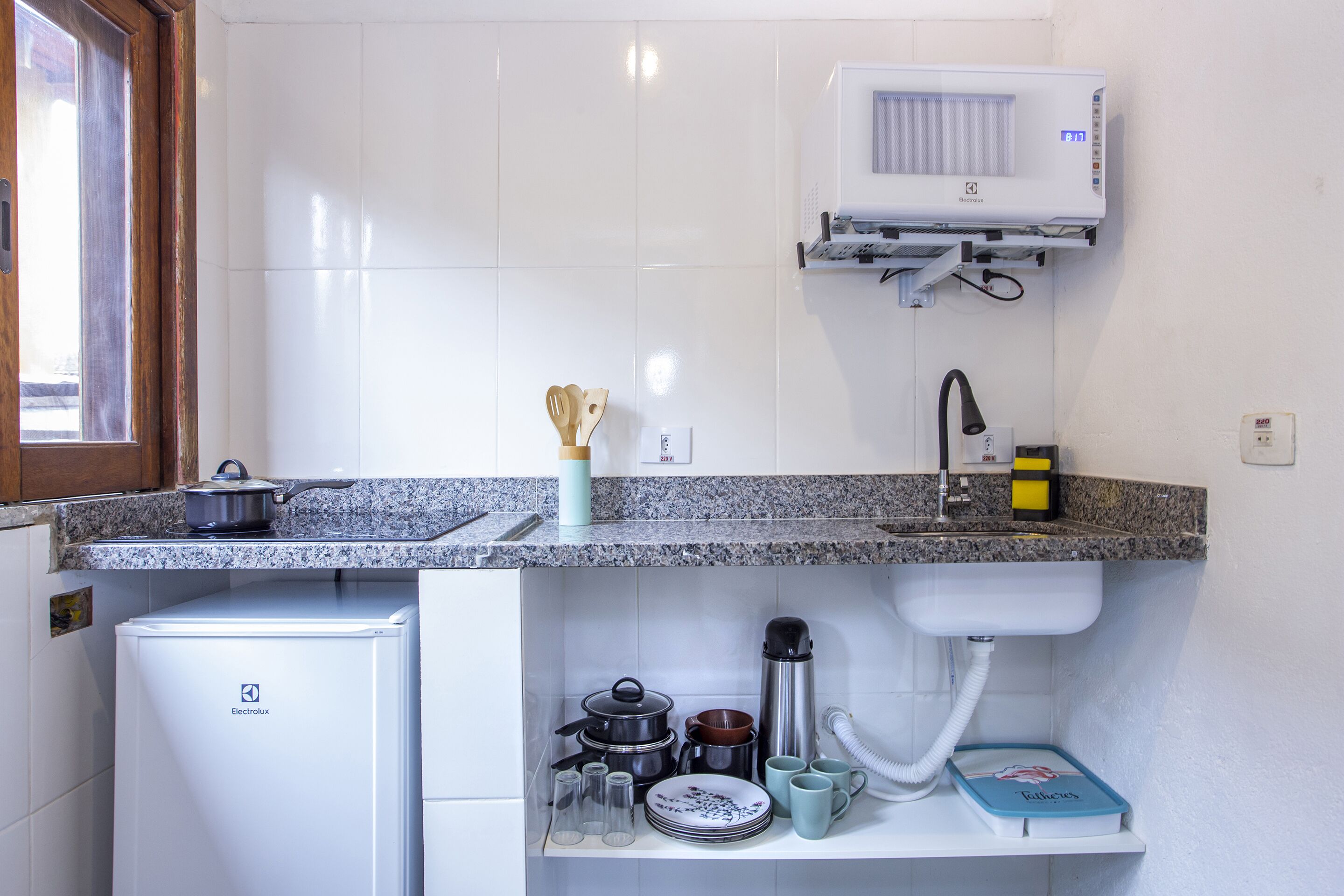 Una cocina compacta con azulejos blancos, microondas, un refrigerador pequeño y diversos utensilios de cocina.