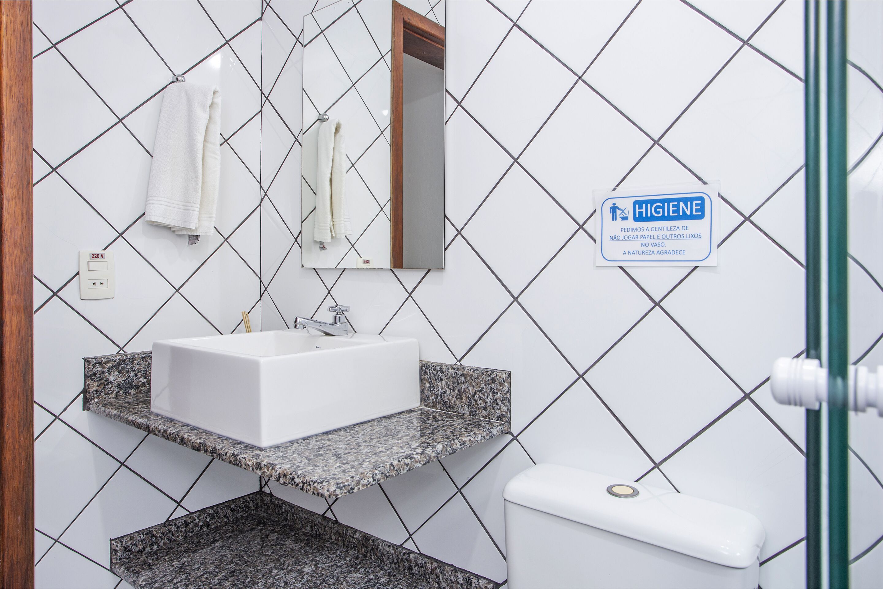 Een badkamer met een wit wastafel, spiegel en toilet. Er hangt een hygiènesymbool op de muur en een handdoekhouder met handdoeken.