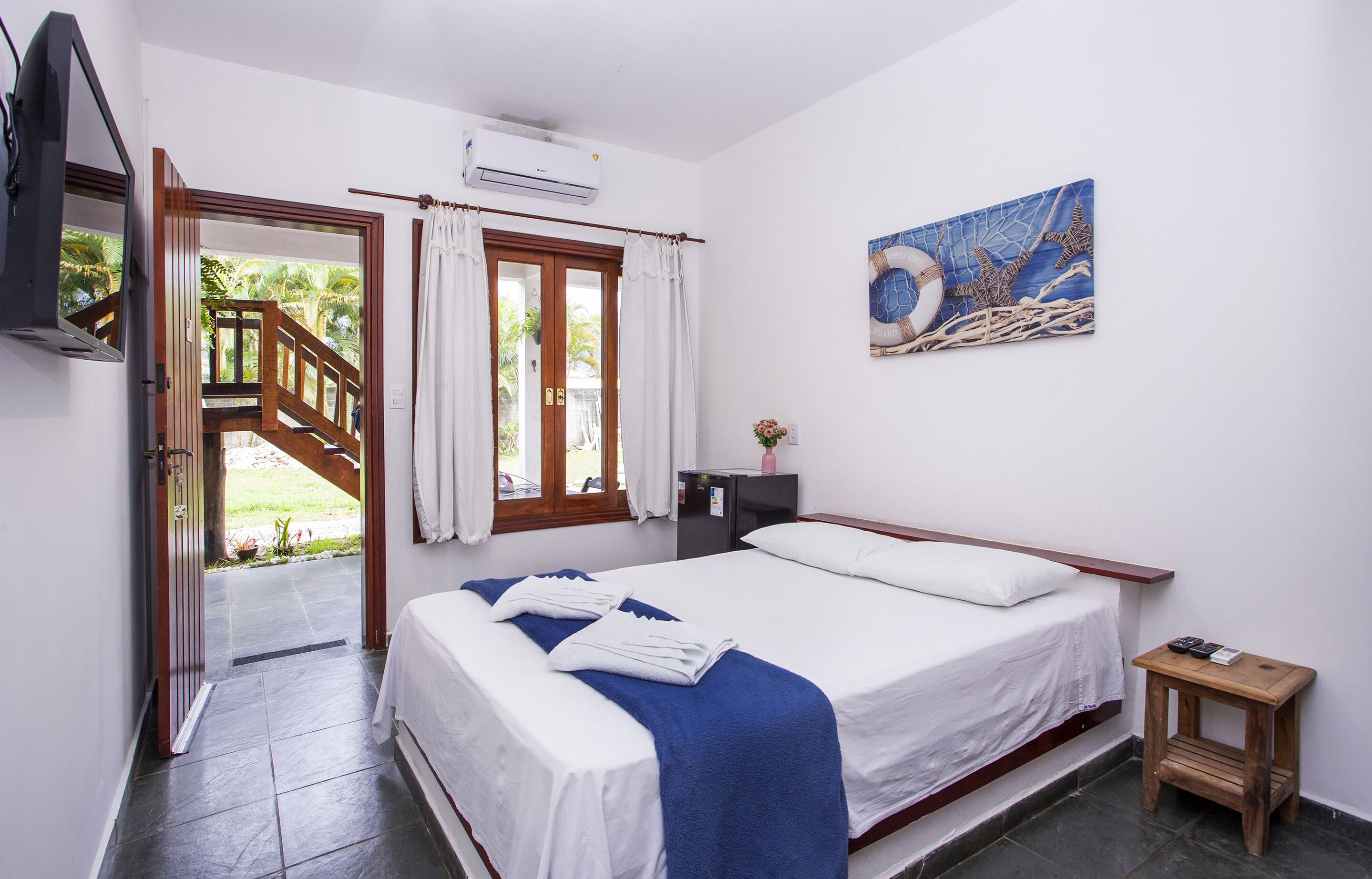 A bedroom with a bed, a small wooden nightstand, a painting, an air conditioning unit, a TV, and a staircase leading to an outdoor area.