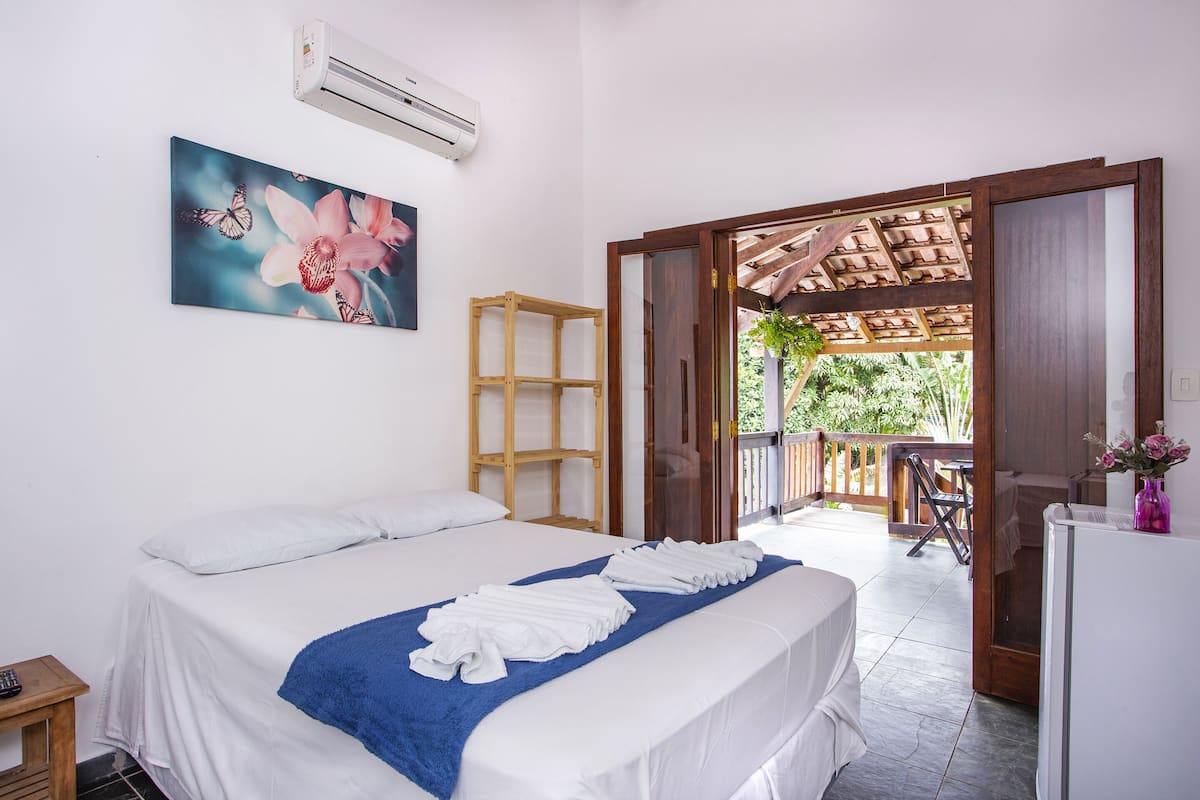 A hotel room with a bed, a blue towel, a wooden shelf, an air conditioner, and a balcony with a table and chairs.