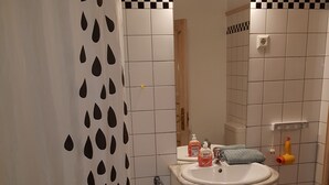 A bathroom with a sink, mirror, and shower. There are toiletries on the sink and a towel rack. - Hansa Apartments Hamburg - Apartment 1B (Wandsbek)