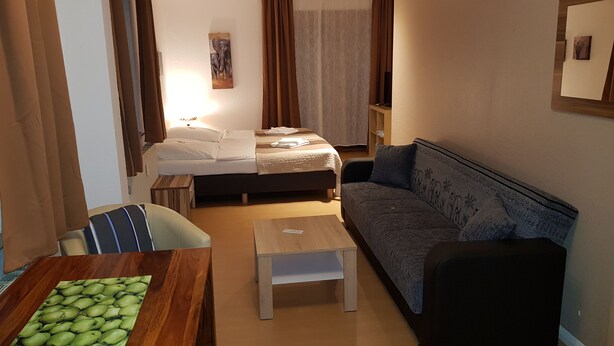 A compact hotel room with a bed, a sofa, a small table, and a dining table with a fruit placemat. - Hansa Apartments Hamburg - Apartment 1B (Wandsbek)