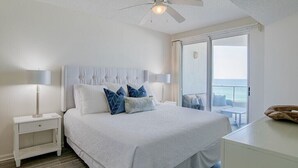 A bedroom with a large bed, two bedside tables, a ceiling fan, and a view of the ocean through a sliding glass door. - Silver Beach 402 | Updated, Direct Beach Front, Pet Friendly! (Orange Beach)