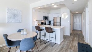 A modern kitchen with white cabinets, a stainless steel refrigerator, and a microwave. There is a dining area with a round table and bar stools. - Silver Beach 402 | Updated, Direct Beach Front, Pet Friendly! (Orange Beach)
