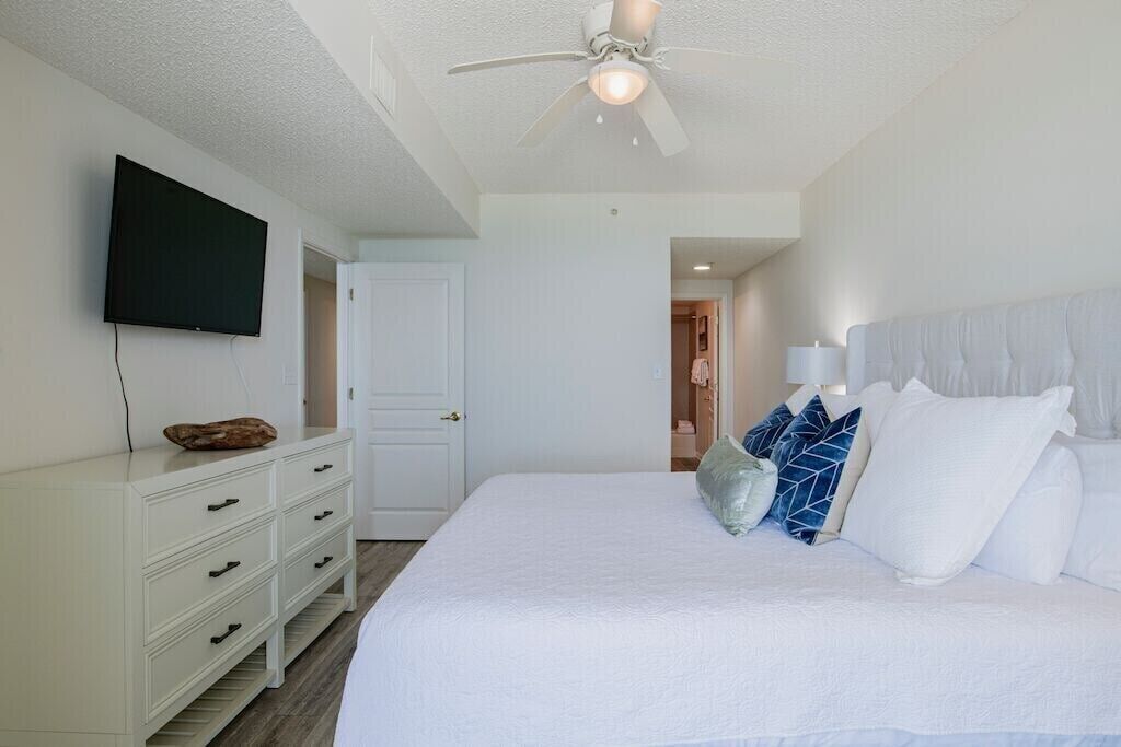 A bedroom with a bed, a ceiling fan, a TV mounted on the wall, and a white dresser.