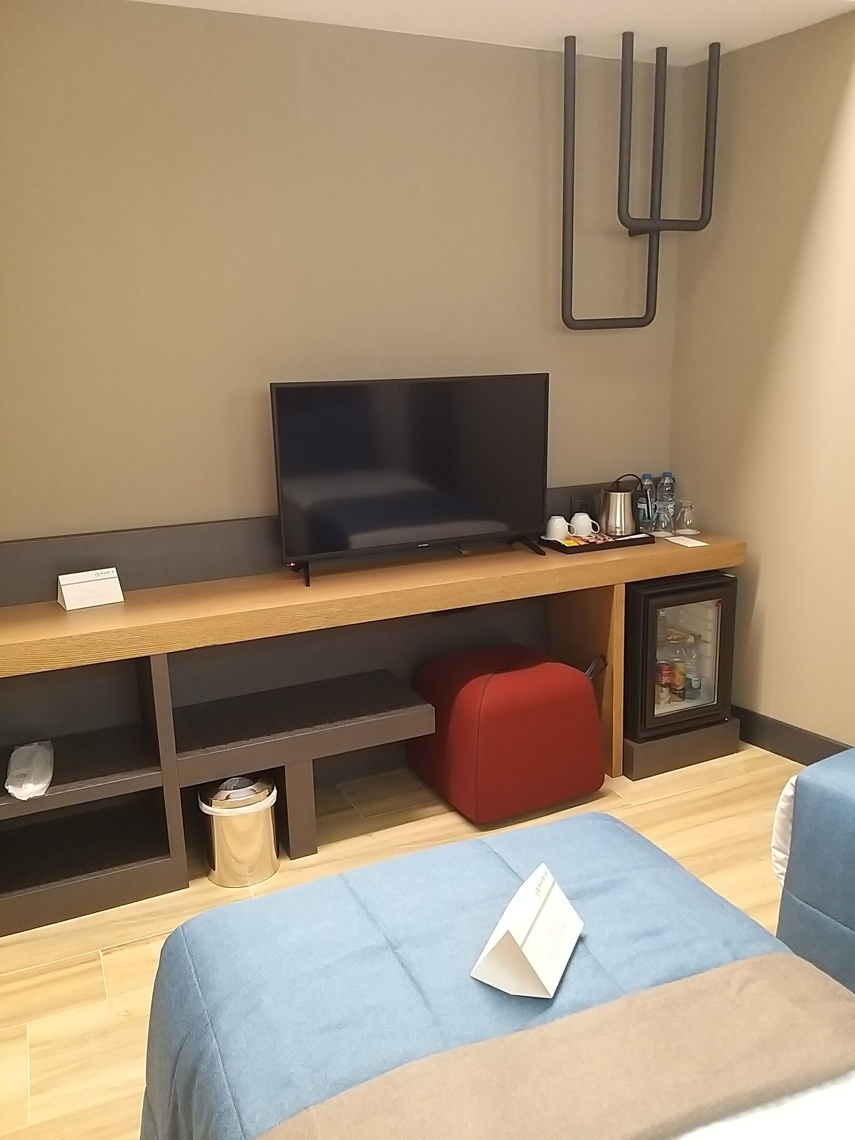 A modern living room with a flat-screen TV, a wooden coffee table, a red ottoman, and a small refrigerator.