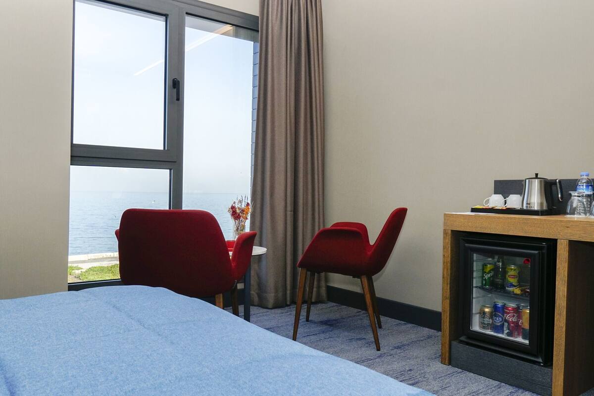 A hotel room with a bed, a desk with a coffee maker, a refrigerator with beverages, a window with a view of the sea, and two red chairs.