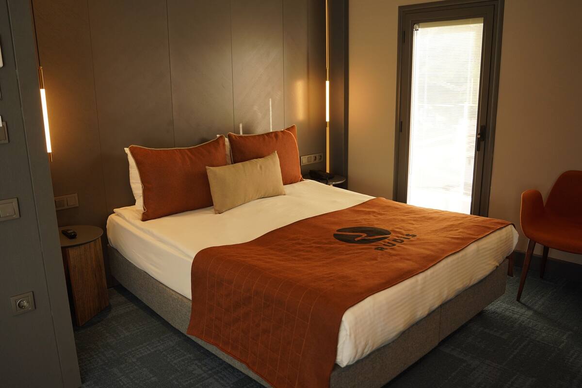 A neatly made bed with a brown blanket and pillows, a bedside table with a lamp, and a chair in a room with a window and a door.