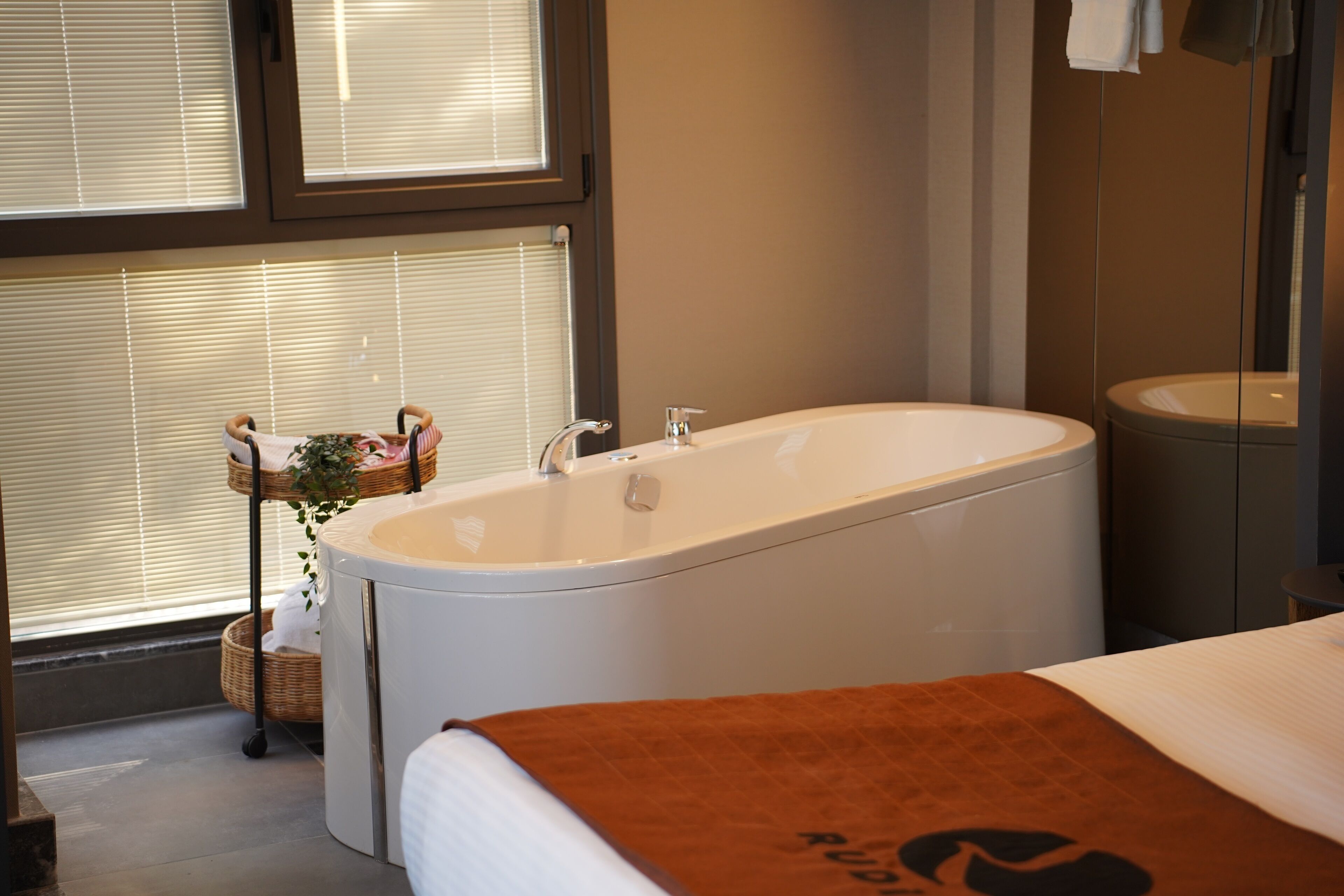 A modern bathroom with a freestanding bathtub, a large mirror, and a window with blinds.