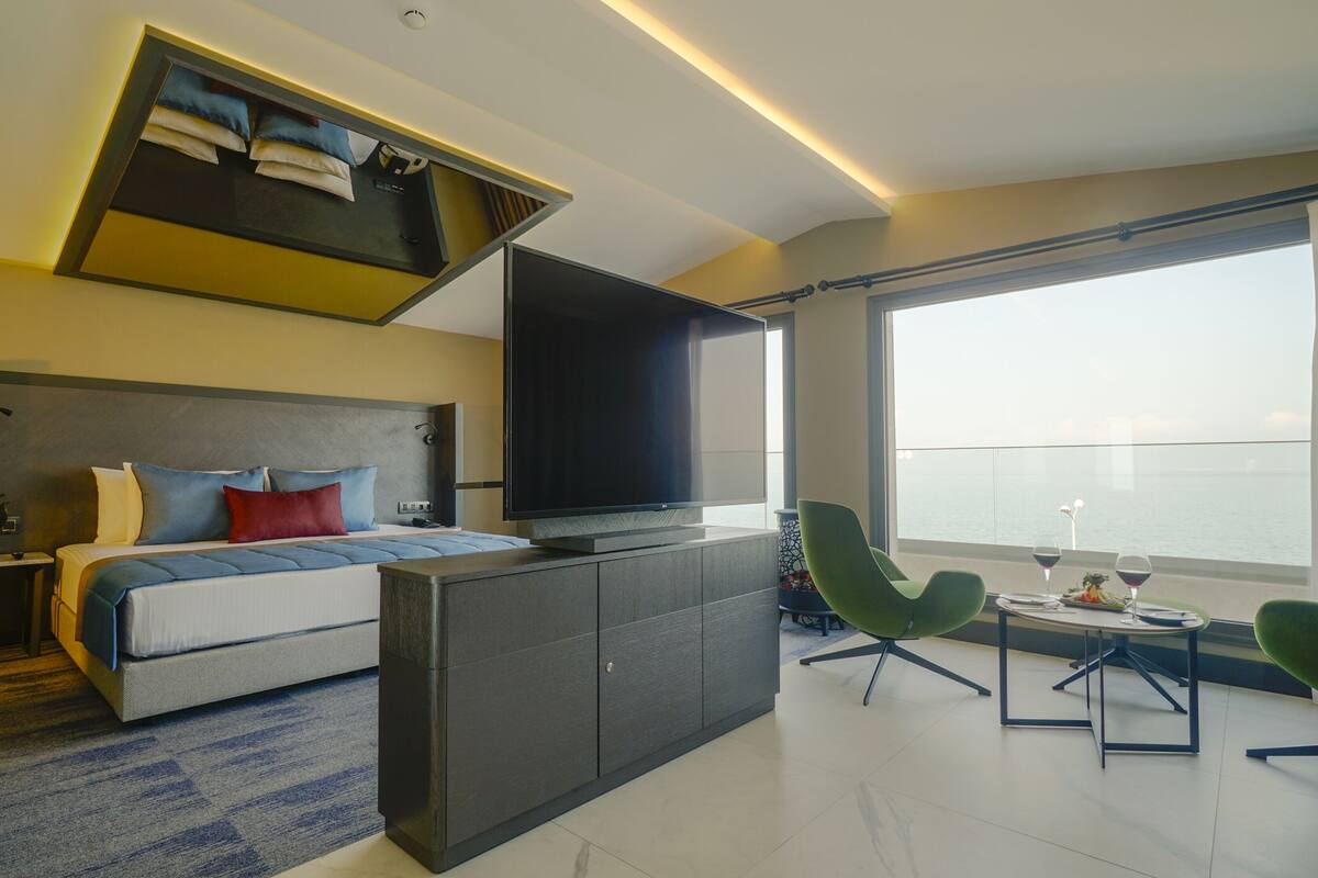 A modern hotel room with a bed, a flat-screen TV, a dining area with chairs, and a large window offering a view of the sea.