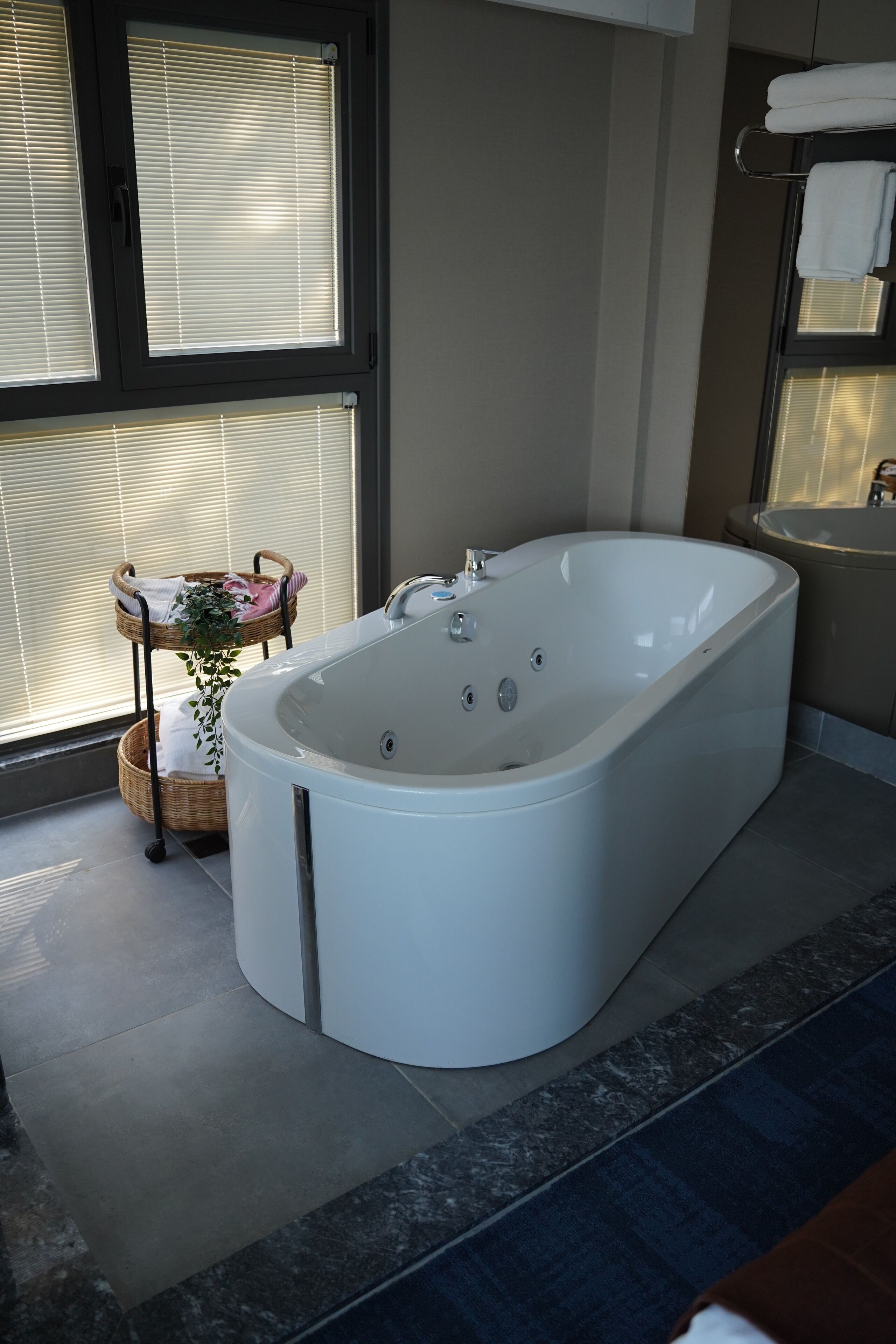 A modern bathroom with a freestanding bathtub, a wicker basket with towels, and a window with blinds.