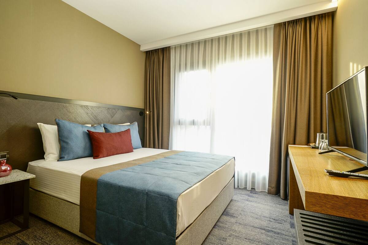 A hotel room with a bed, a desk with a TV, a nightstand, and a window with curtains.