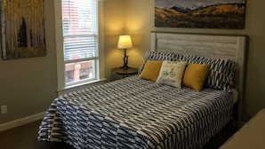 A bedroom with a bed, a nightstand, a lamp, a window with blinds, and a painting on the wall. - Loft Apartment - Manitou (Manitou Springs)