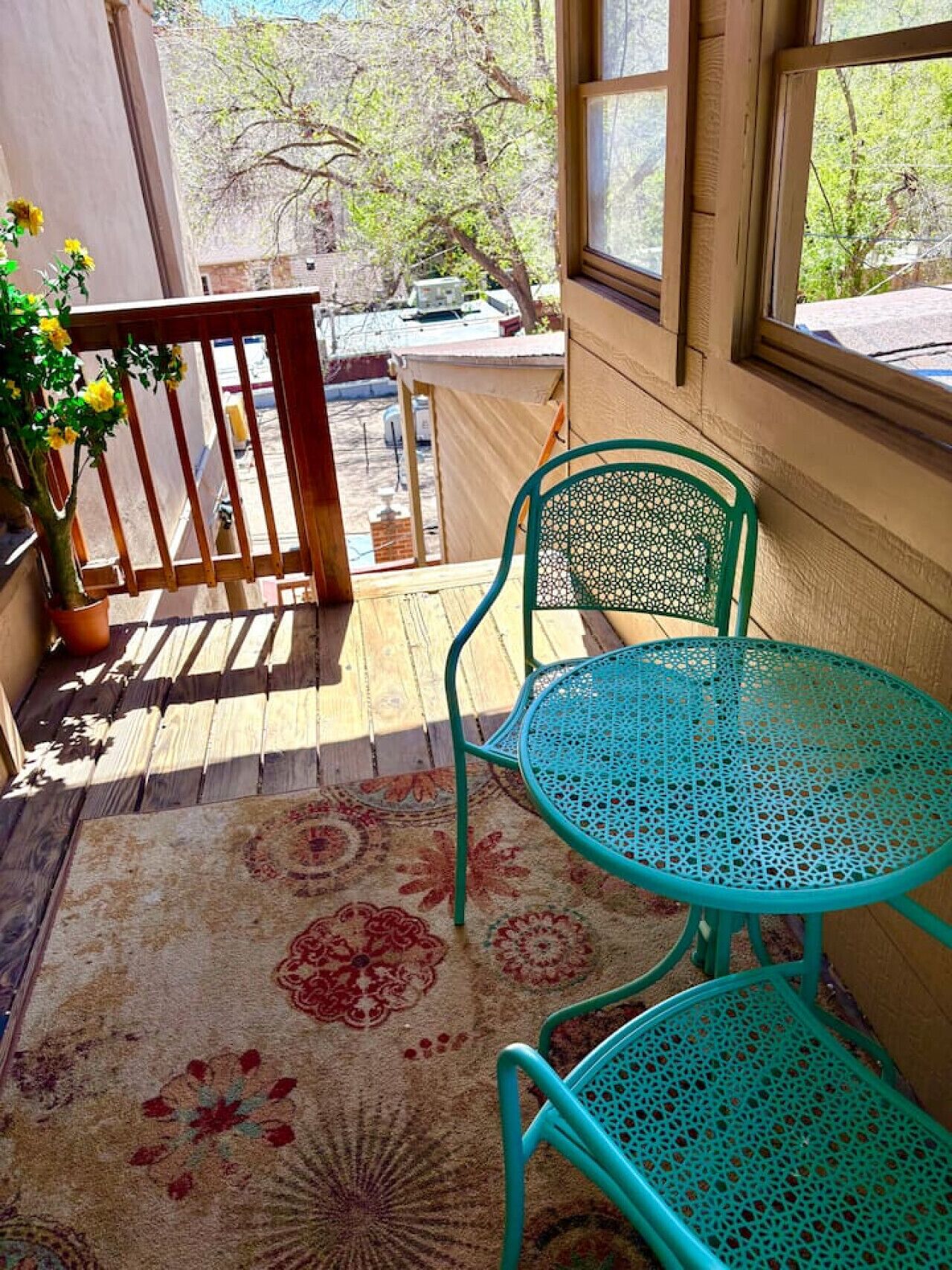 A wooden deck with a turquoise metal table and chairs, a potted plant, and a patterned rug.