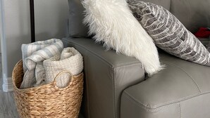 Un sofá de cuero gris con un almohadón blanco mullido y un almohadón estampado, una cesta de mimbre con mantas, y una lámpara de pie con pantalla de vidrio transparente.