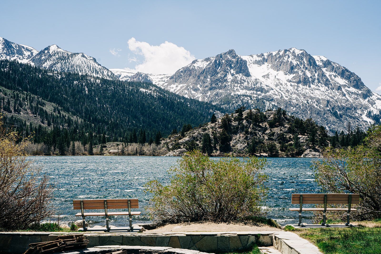 A lakeside view with two wooden benches facing the water, surrounded by mountains and trees.
